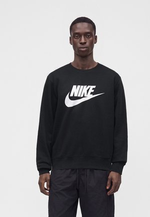 Άνδρας που φοράει μαύρο φούτερ Nike με λευκό λογότυπο και μαύρο παντελόνι, στέκεται μπροστά σε απλό φωτεινό φόντο.