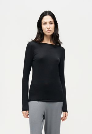 Une femme porte un haut noir à manches longues et un pantalon gris sur un fond clair et uni.