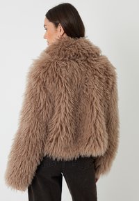 Brun faux fur jakke med et struktureret, voluminøst design, cropped pasform og brede ærmer. Har en høj krave og bæres over sorte jeans.