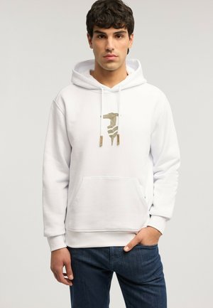 Junger Mann trägt einen weißen Hoodie mit goldenem grafischem Logo, dunkelblaue Jeans, steht vor einem schlichten hellen Hintergrund.