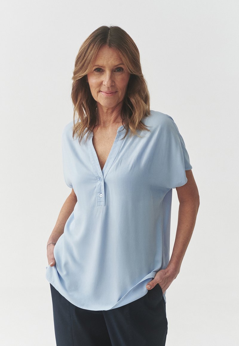 Mujer de mediana edad con cabello castaño claro hasta los hombros que lleva una blusa azul claro y pantalones oscuros, de pie con las manos en los bolsillos frente a un fondo blanco.