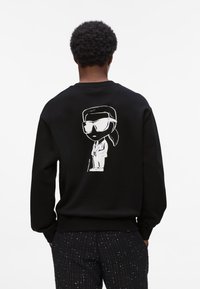Person mit kurzem, lockigem Haar trägt einen schwarzen Pullover mit einem weißen Comic-Charakter, der Sonnenbrillen, einen Anzug und einen Pferdeschwanz auf dem Rücken hat.