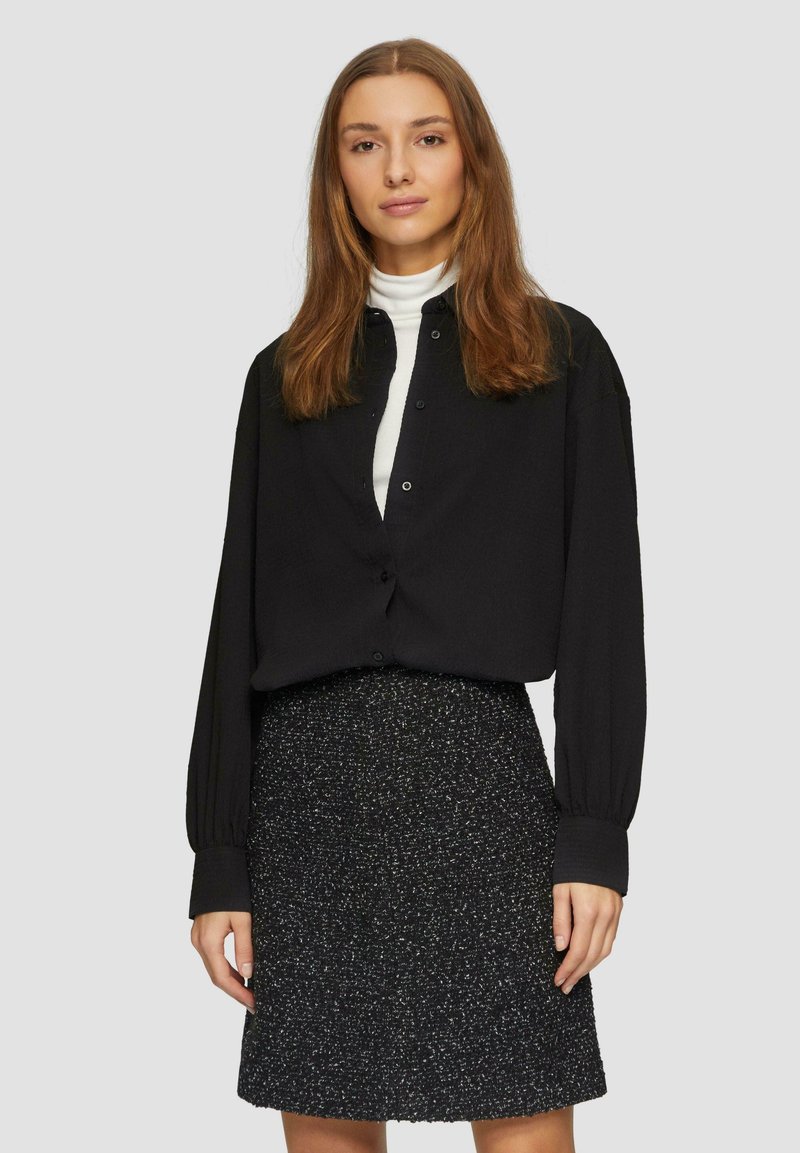 Zwarte button-up blouse met poffende mouwen, gecombineerd met een gestructureerde zwarte rok met een subtiele zilveren glinstering. Effen witte binnenlaag zichtbaar.
