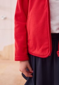 Giacca in pile rosso con chiusura a zip e tessuto strutturato, abbinata a una gonna scura. Mano visibile appoggiata in vita, cursore della zip liscio.