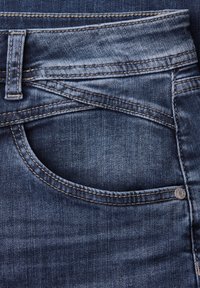 Nærbillede af mørkeblå denimjeanslomme med detaljeret syning, bæltestrop og metalnitte på tekstureret stof.
