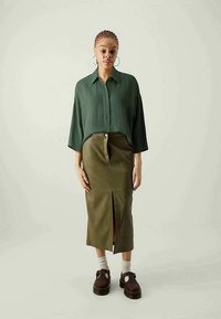 Camisa oversized plissada verde, saia verde-oliva de cintura alta com fenda frontal, sapatos de plataforma castanhos, meias brancas, brincos grandes em argola.