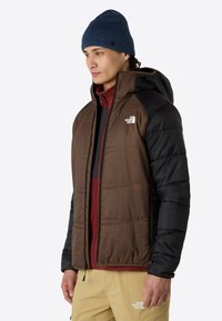 Braune und schwarze isolierte Jacke mit Kapuze, gestepptem Muster, zwei Seitentaschen, über einem weinroten Shirt und beigen Hosen getragen. Blaue Strickmütze.