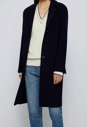 Osoba ubrana w granatowy płaszcz na guziki, pod którym ma biały sweter z dekoltem w serek z czarną lamówką oraz jasnoniebieskie dżinsy, stoi na jasnym tle.