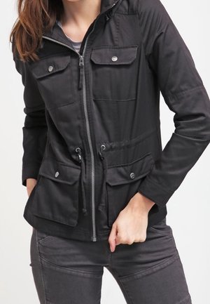 Henkilö, joka pukeutunut mustaan vetoketjulliseen takkiin, jossa on neljä etutaskua läpällä ja vyötäröllä kiristysnauhat, yhdistettynä tummanharmaisiin housuihin.