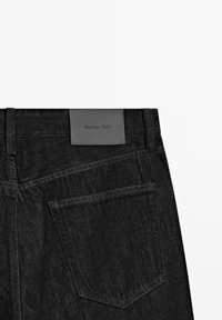 Sorte denimbukser med en lædermærkat med teksten "Massimo Dutti." Inkluderer kontrasterende syning og en baglomme med et buet design.