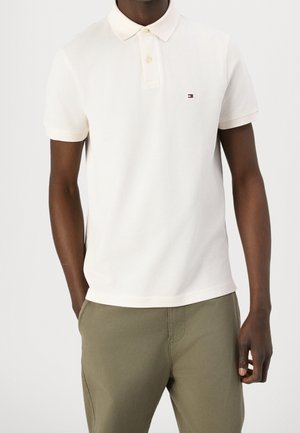 Mann trägt ein weißes kurzärmeliges Poloshirt mit kleinem Tommy Hilfiger-Logo, kombiniert mit olivgrünen Hosen.