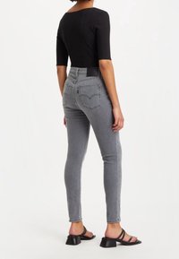 Jeans de tiro alto gris de denim, con un diseño ajustado, dos bolsillos traseros y dobladillos deshilachados, combinados con una blusa negra ajustada y sandalias.