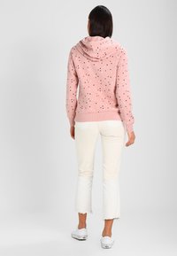 Felpa rosa chiaro con piccoli punti neri e bianchi, dotata di cappuccio con cordino, polsini a coste e una vestibilità comoda, abbinata a jeans bianchi a zampa d'elefante.