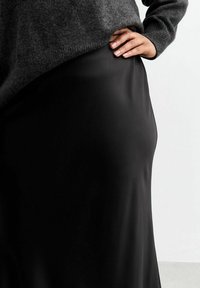 Jupe noire en satin avec une texture lisse, tombant doucement. La taille est recouverte par un pull en tricot gris reposant juste au-dessus.