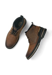 Des bottines en cuir marron présentent une finition lisse, des accents noirs, un bout rond et une semelle en caoutchouc texturée avec des détails de couture.