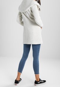 Abrigo largo de punto gris claro con una gran capucha, tejido texturizado, dos bolsillos laterales, combinado con jeans ajustados azules y zapatillas negras.