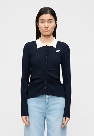 Marineblauw geribd vest met een witte kraag, knoopsluiting aan de voorkant en geplooide details bij de taille. Heeft een logo aan de linkerkant.