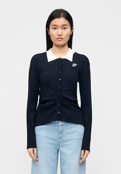 Cardigan côtelé bleu marine avec un col blanc, une fermeture à boutons sur le devant et un détail froncé à la taille. Dispose d'un logo sur le côté gauche.