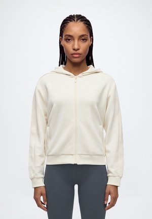 Une femme aux cheveux tressés se tient face à l’objectif, vêtue d’un sweat à capuche crème zippé et d’un legging gris foncé, devant un fond clair uni.