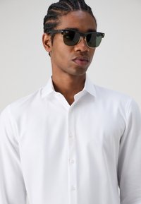 Weißes, langärmliges Button-up-Hemd mit klassischem Kragen, getragen mit schwarzen Sonnenbrillen mit grünen Linsen und goldenen Akzenten.