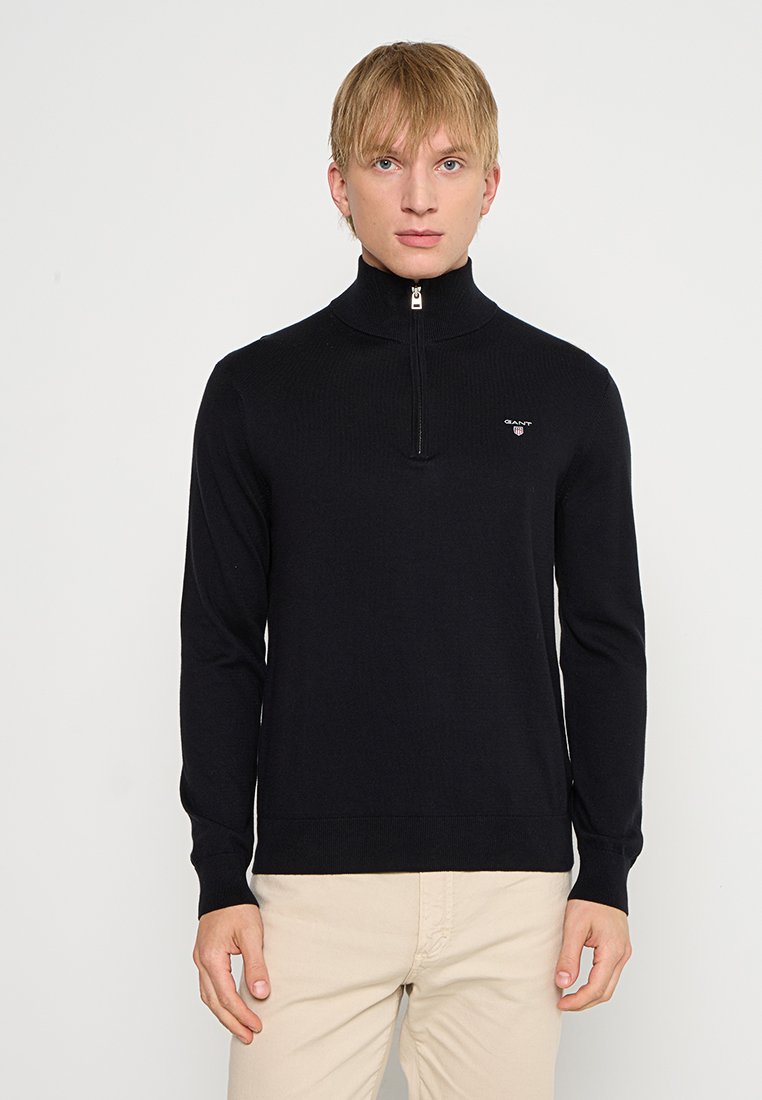 Maglione nero lavorato a maglia con colletto rialzato e chiusura a mezza zip. Presenta un piccolo logo sul petto e polsini e orlo aderenti.