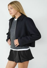 Schwarze, kurz geschnittene Jacke mit Kragen, aus leichtem Stoff, mit langen Ärmeln. Grauer Pullover darunter getragen, kombiniert mit einem schwarzen Faltenrock.