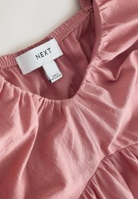 Encolure de vêtement en coton rose avec bord élastique froncé et étiquette de marque blanche indiquant "NEXT" et étiquette pays "Made in Bangladesh".