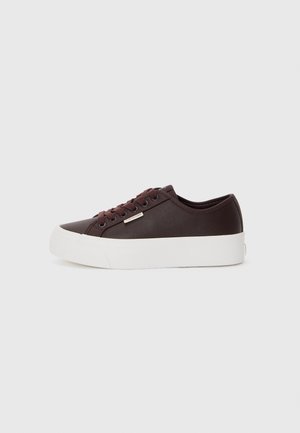 Ciemnobrązowy sneaker z niską cholewką, pasującymi sznurówkami, grubą białą gumową podeszwą i małą metalową metką z logo z boku, pokazany na jednolitym tle.