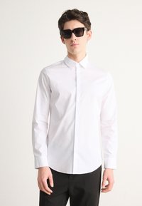 White button-up shirt with a pointed collar, long sleeves, and smooth texture; model wearing dark sunglasses and black pants.