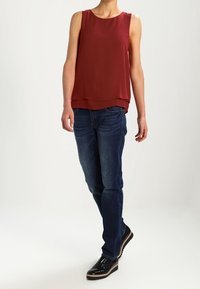 Bordeauxfarvet ærmeløs top med lagdelt kant, parret med blå denimjeans og skinnende sorte sko. Enkel, moderne skæring uden mønstre eller accenter.