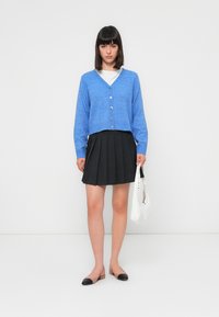 Hellblauer, kürzer geschnittener Cardigan aus weichem Strickstoff, mit V-Ausschnitt und fünf Knopfverschlüssen. Kombiniert mit einem schwarzen, plissierten Rock.