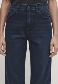 Jeans in denim blu scuro con vita alta, cinque tasche, passanti per cintura e un classico bottone. Texture liscia con sottili dettagli di cucitura.