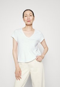 Balts īsām piedurknēm T-krekls ar V veida kakla izgriezumu, piedāvājot brīvu piegriezumu un nedaudz nodilušus malas. Kombinēts ar gaiši bēšiem biksēm.