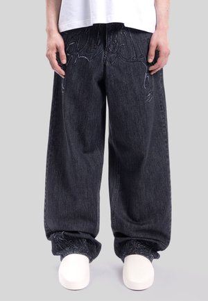 Persona que lleva vaqueros sueltos de denim oscuro con discretos diseños bordados cerca de los bolsillos y los puños, combinados con zapatos blancos lisos y una camisa blanca.