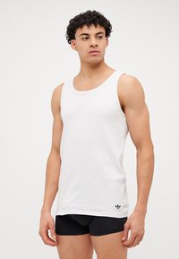 Λευκή αμάνικη μπλούζα φτιαγμένη από απαλό βαμβακερό ύφασμα; διαθέτει στρογγυλή λαιμόκοψη και διακριτική μαύρη λογότυπη πινελιά στο τελείωμα.