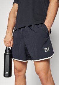 Marine treningsshorts med hvit kant, laget av lett materiale. Har et Nike-logo-patch. En svart Nike-vannflaske holdes i hånden.