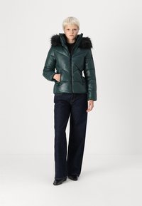 Green puffer jacket with a glossy finish, featuring a removable black faux fur hood, zip front, and diagonal quilted pattern. Paired with dark jeans.