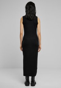 Robe maxi sans manches noire avec une silhouette ajustée, une texture lisse et un design long jusqu'au sol, associée à des bottes noires épaisses.