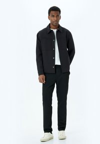 Zwarte buitenjas met een kraag, twee borstzakken en knoopsluiting, gecombineerd met zwarte broek en witte sneakers.