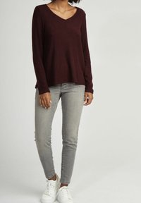 Burgunderfarbendes Langarmshirt mit V-Ausschnitt, kombiniert mit hellgrauen, figurbetonten Jeans und weißen Sneakers. Es hat eine lässige Passform und seitliche Schlitze.