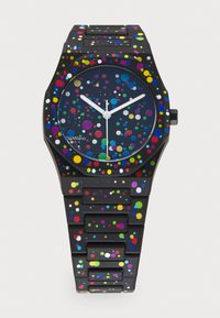 Montre en plastique noir avec un cadran rond, présentant un design moucheté multicolore sur le cadran et le bracelet, avec des aiguilles des heures et des minutes blanches.