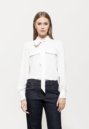Weiße Bluse mit einer Krawatten-Halslinie und zwei Brusttaschen, kombiniert mit dunkelblauen Jeans aus Denim mit sichtbaren Nähten und Taschen.