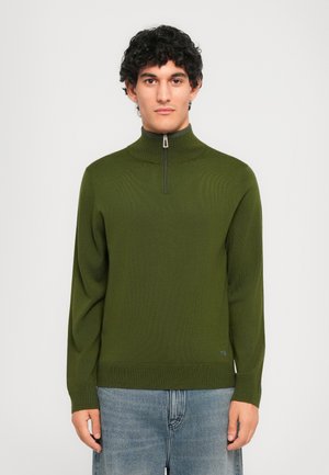 Man with curly black hair wearing an olive green half-zip sweater and blue jeans, standing against a plain light background.