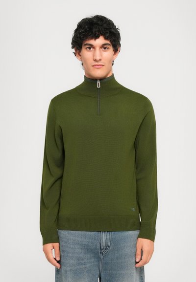 Uomo con capelli neri ricci, che indossa un maglione olive green a mezza zip e jeans blu, in piedi di fronte a uno sfondo chiaro e semplice.