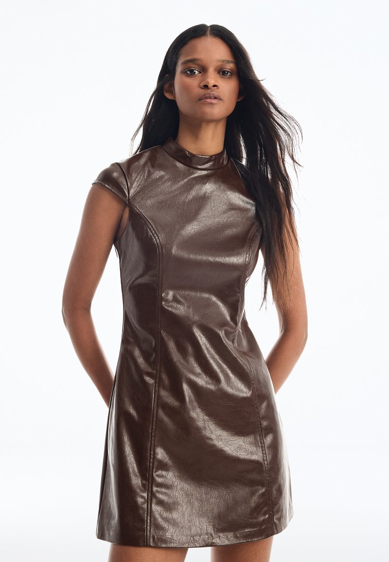 Jeune femme aux longs cheveux foncés portant une mini-robe en cuir marron sans manches, debout devant un fond blanc uni.