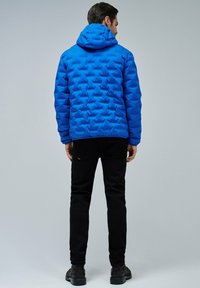 Hombre de pie de espaldas, vistiendo una chaqueta acolchada con capucha de color azul brillante, pantalones negros y botas negras, ante un fondo sencillo.