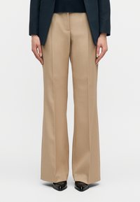 Άτομο που φοράει μπεζ παντελόνια ραμμένα στα μέτρα, μαύρα μυτερά παπούτσια και σκούρο σακάκι, στέκεται μπροστά σε απλό φόντο.