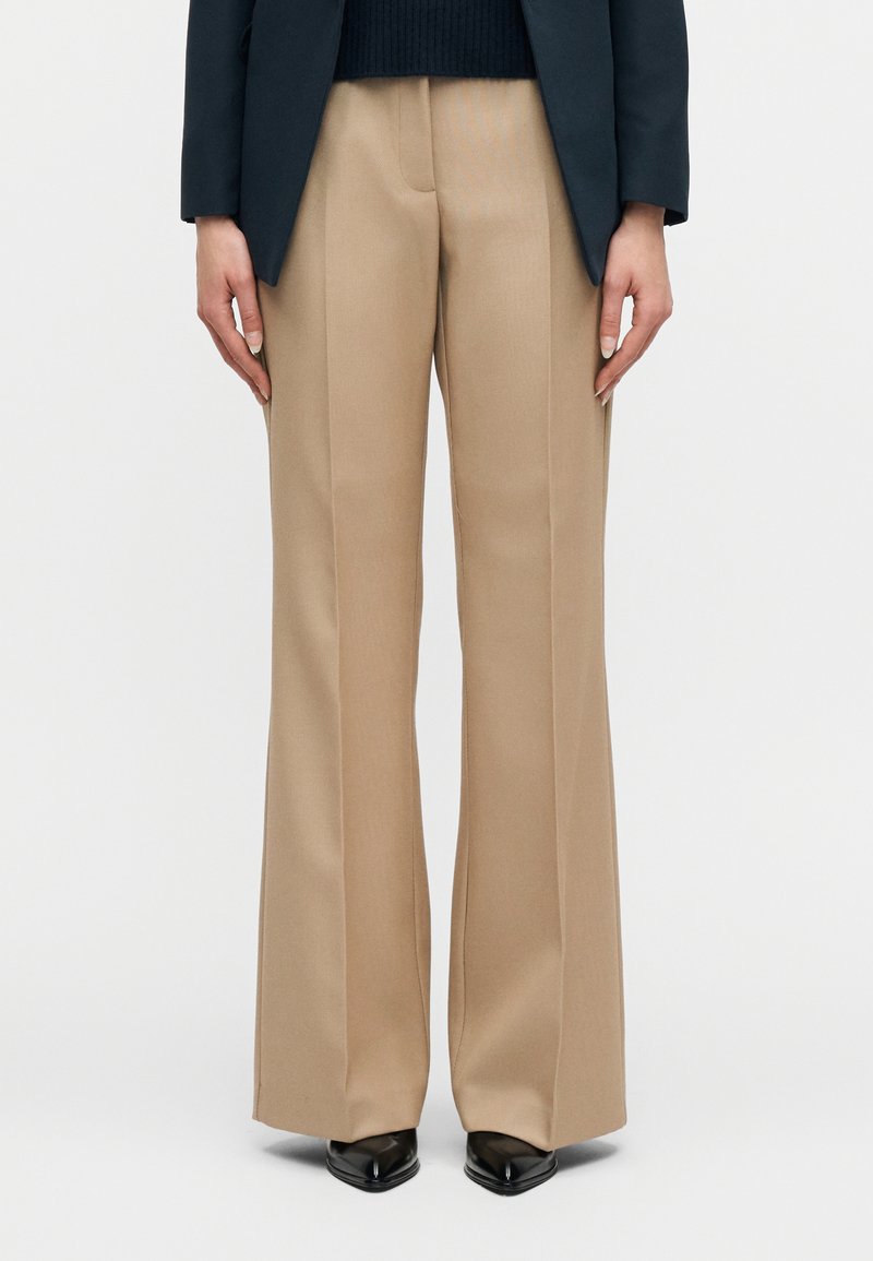 Άτομο που φοράει μπεζ παντελόνια ραμμένα στα μέτρα, μαύρα μυτερά παπούτσια και σκούρο σακάκι, στέκεται μπροστά σε απλό φόντο.