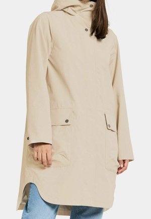 Vrouw draagt een beige regenjas met capuchon en knoopsluitende klepzakken, staat met zichtbare handen, gecombineerd met blauwe denim jeans.
