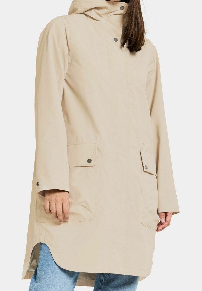 Femme portant un imperméable à capuche beige avec poches à rabat boutonnées, debout les mains visibles, assorti à un jean denim bleu.